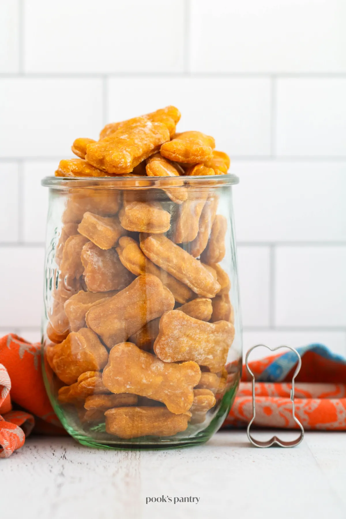 A collection of helpful tools for making soft pumpkin treats, including a glass jar filled with treats and a bone-shaped cookie cutter placed beside it.