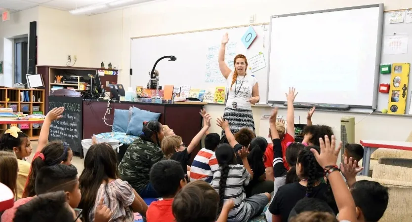 A teacher and several of her students are raising their hands in a Phoenix-area classroom during a group lesson.