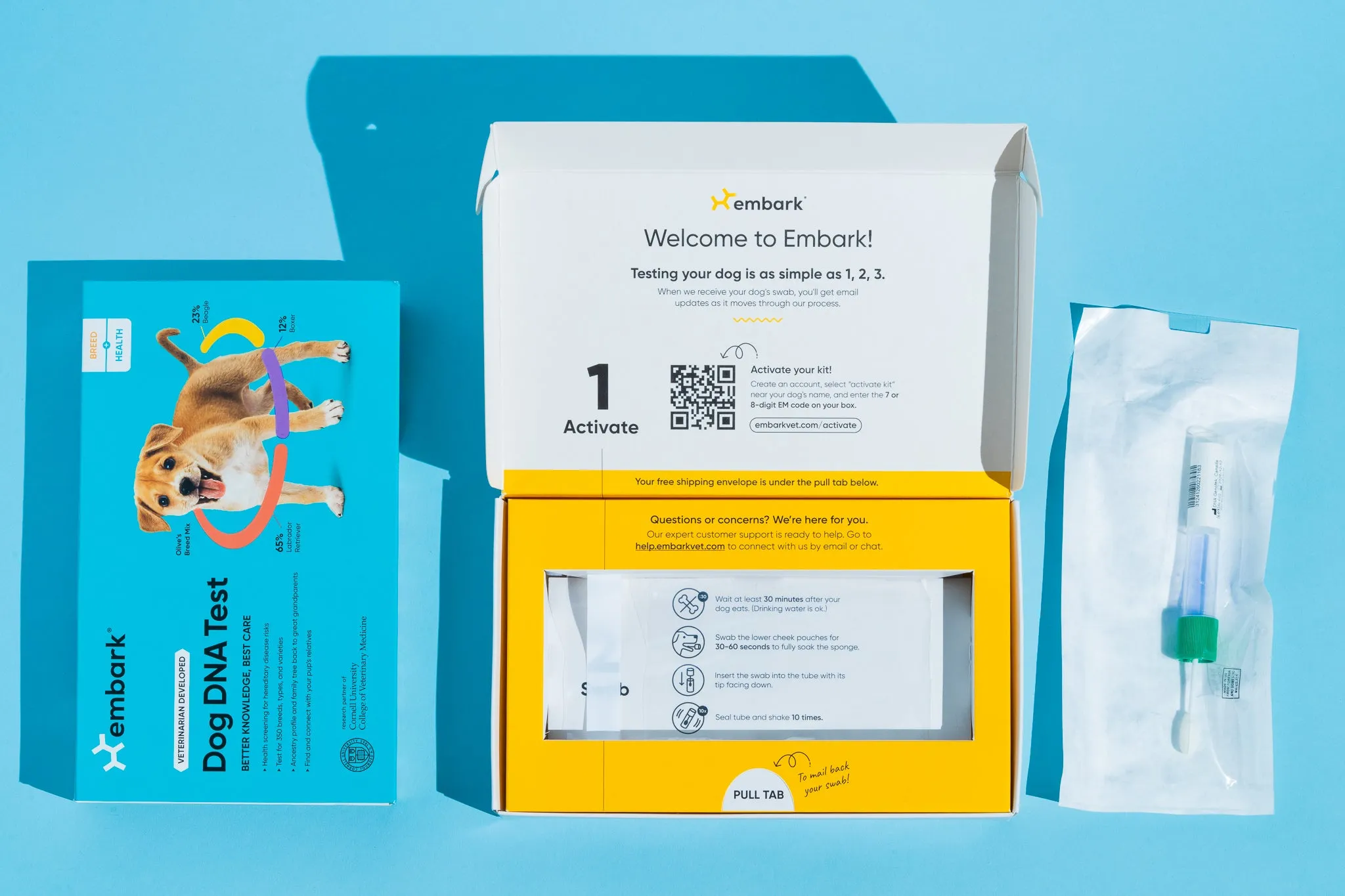 An Embark Dog DNA Test kit displayed showing the blue box, the opened yellow test kit with instructions, and a sealed swap.
