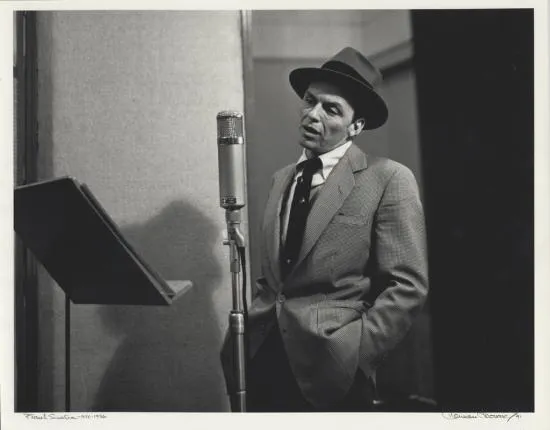 Frank Sinatra in hat and tie stands at microphone with music stand in front of him, mouth slightly open