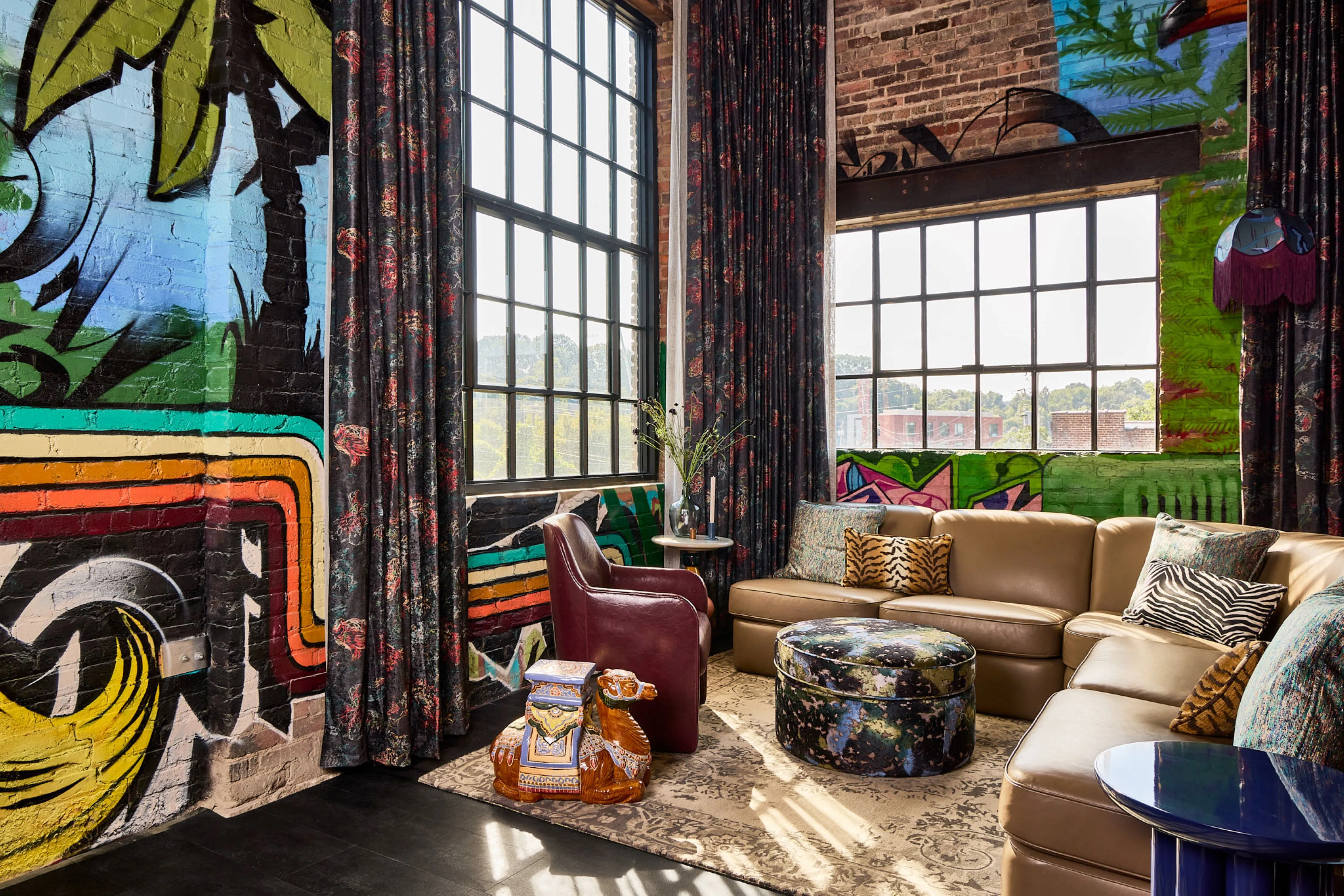 The Radical Asheville, Tapestry Collection by Hilton - Lobby with painted brick wall and leather chair and l-shaped couch with throw pillows next to an ottoman and end tables all sitting on top of a patterned rug