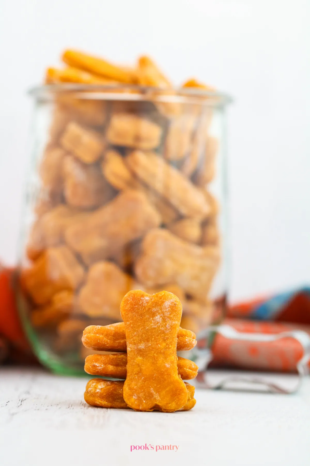A clear glass Weck jar is filled with soft pumpkin dog treats, with more treats piled neatly in front, showcasing their appealing texture and readiness for storage.