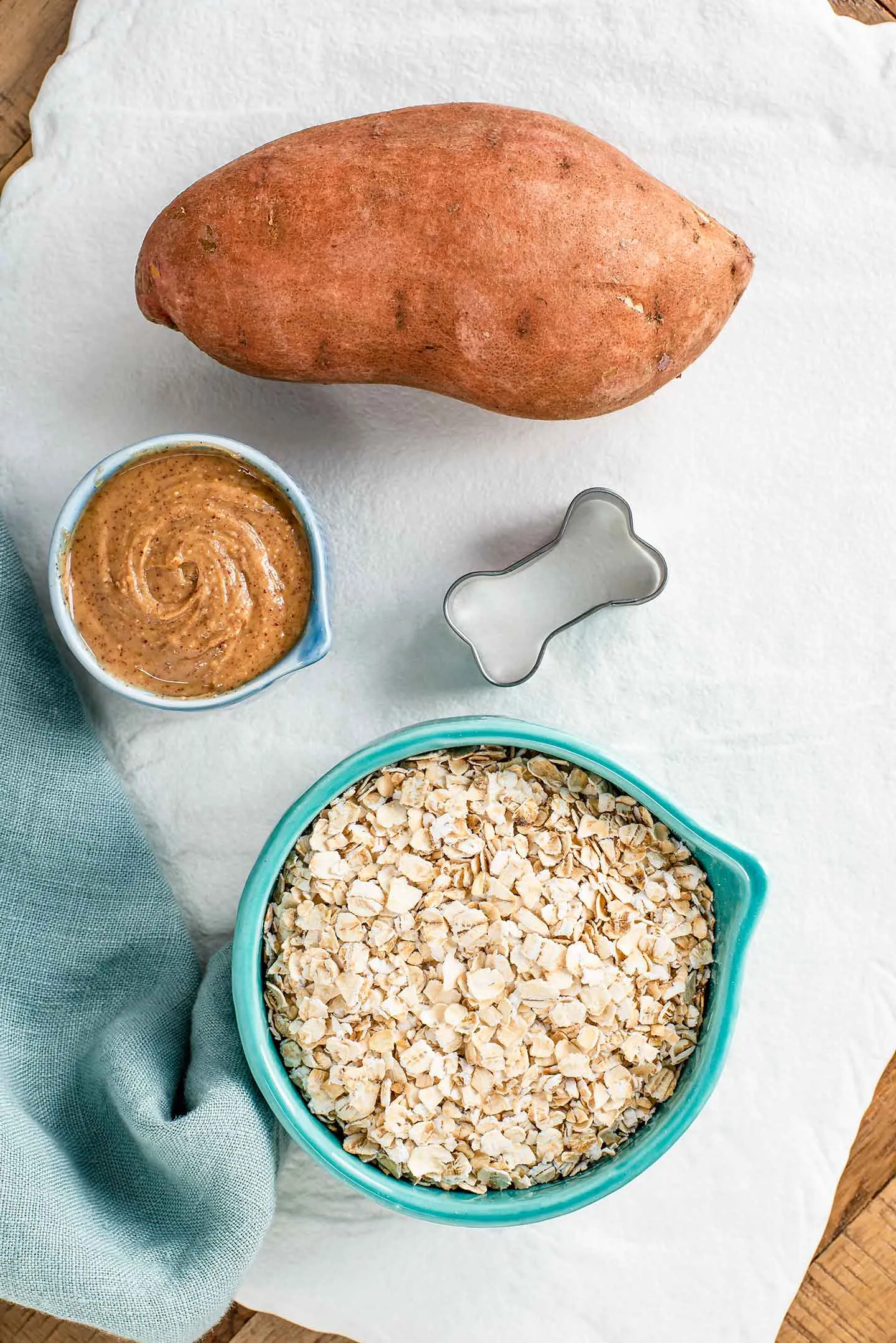 A top-down view of a whole sweet potato, a bowl of peanut butter, a cup of oats, and a bone-shaped biscuit cutter on a white surface.