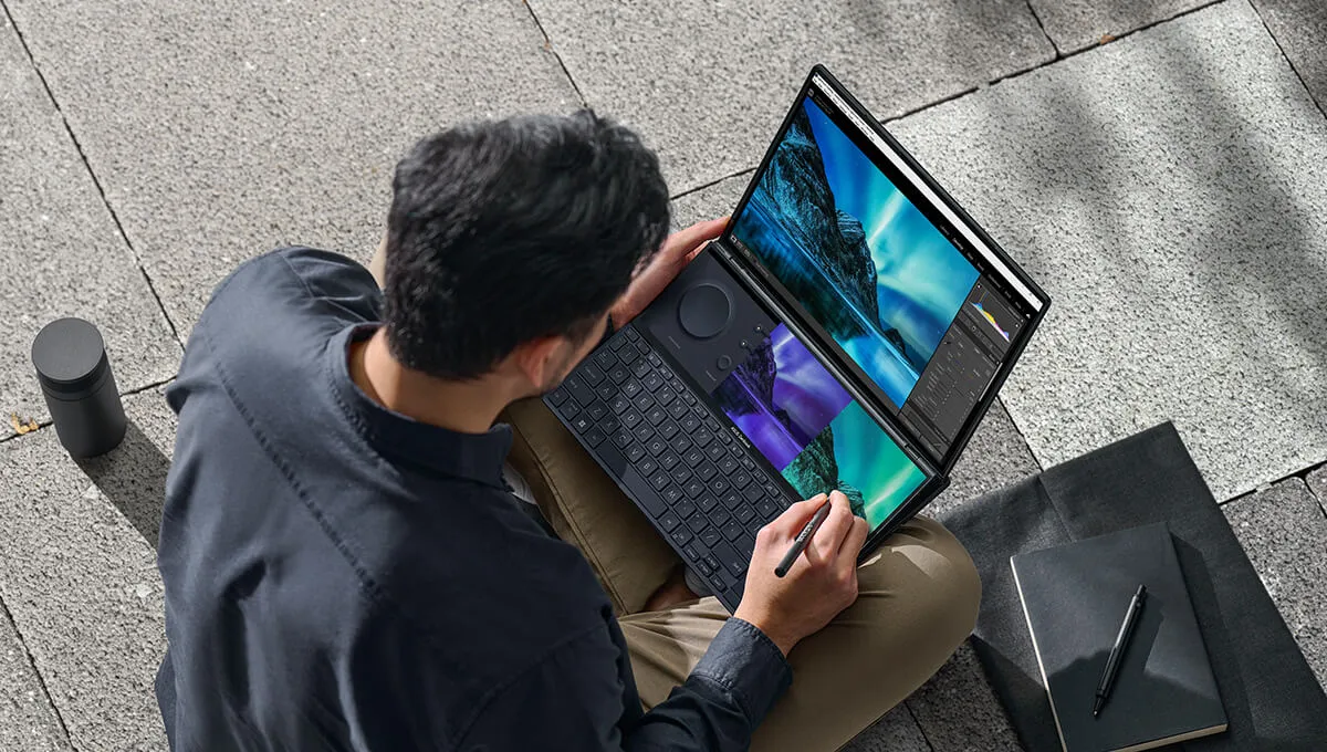 A top view of a young man using a stylus on the ASUS ScreenPad Plus of the ZenBook Pro Duo 14 OLED dual screen laptop while sitting on stairs outdoors