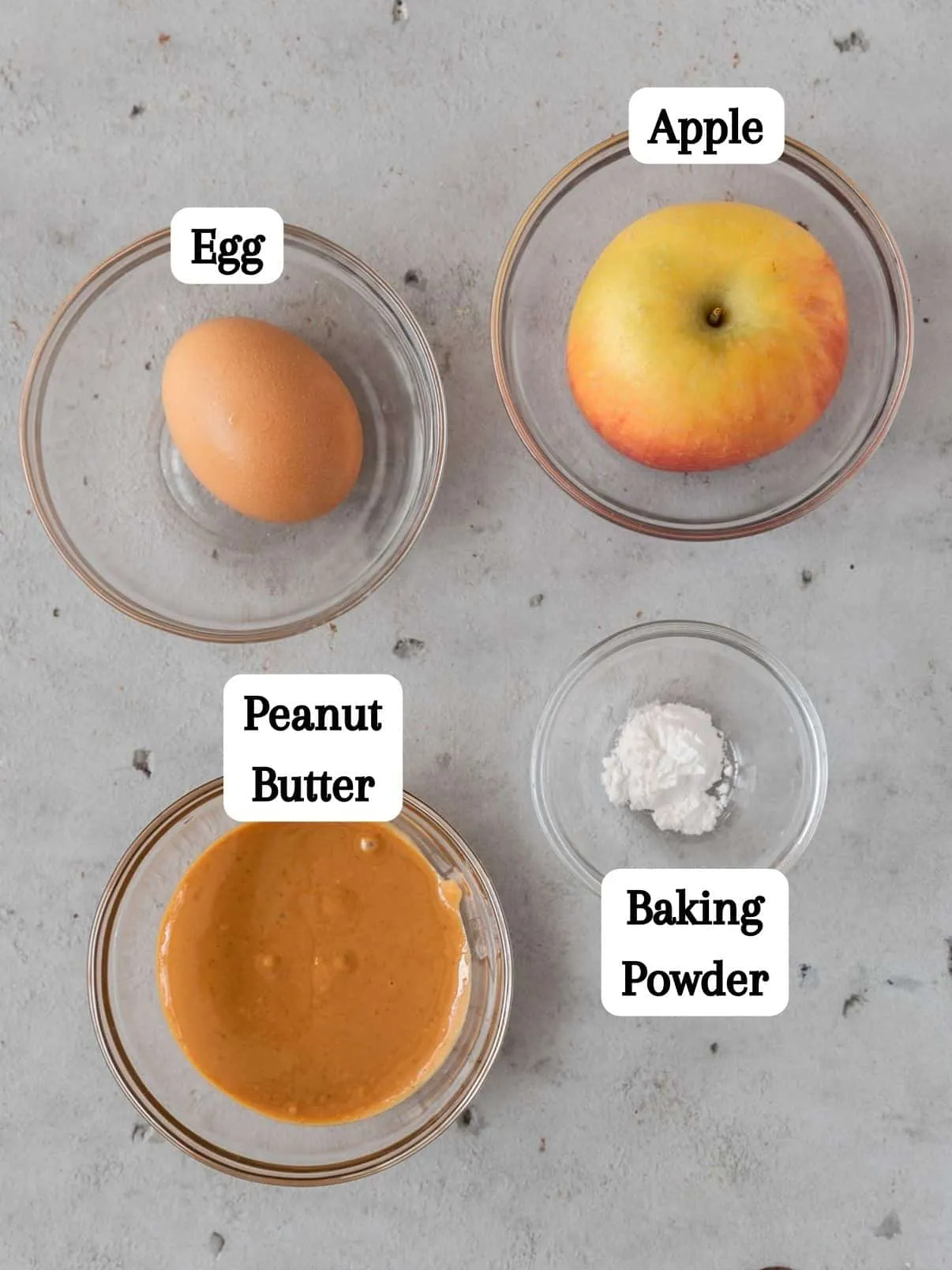 All ingredients for the homemade dog cake recipe, including chopped apple, peanut butter, egg, and baking powder, arranged in glass bowls on a grey background.