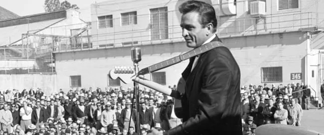 Johnny Cash performing on stage in front of a large crowd of male inmates, many wearing hats and sunglasses.