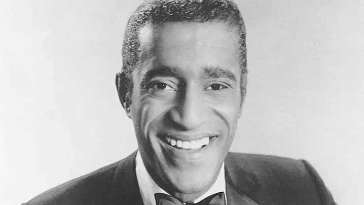 Sammy Davis Jr. with a serious expression, performing on stage with a microphone.