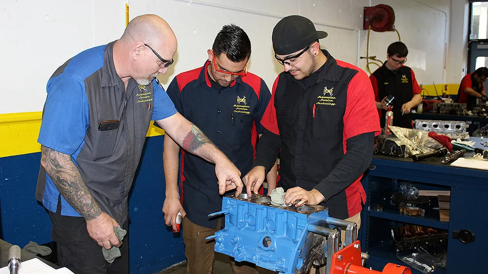 Students observing an engine component with an instructor in an automotive workshop