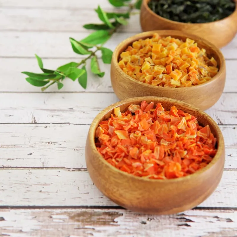 Three rustic wooden bowls filled with various natural, dried ingredients like herbs and vegetables, showcasing wholesome components of pet food.