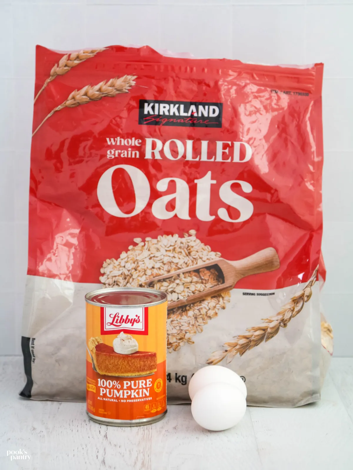 A bag of oatmeal, a can of pumpkin puree, and two eggs on a white background.