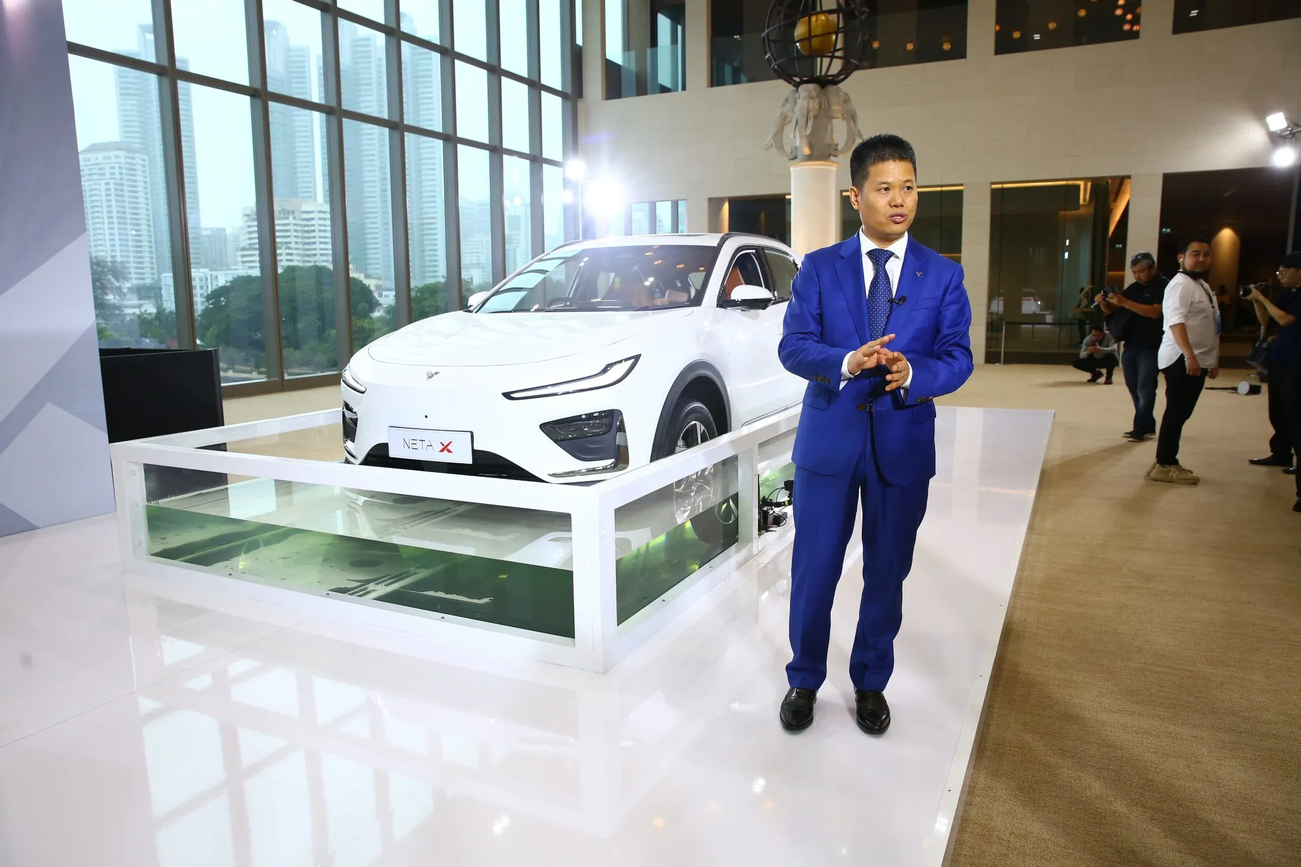 A man in a blue suit stands next to a white car displayed inside a glass showcase, with city buildings visible through large windows in the background.