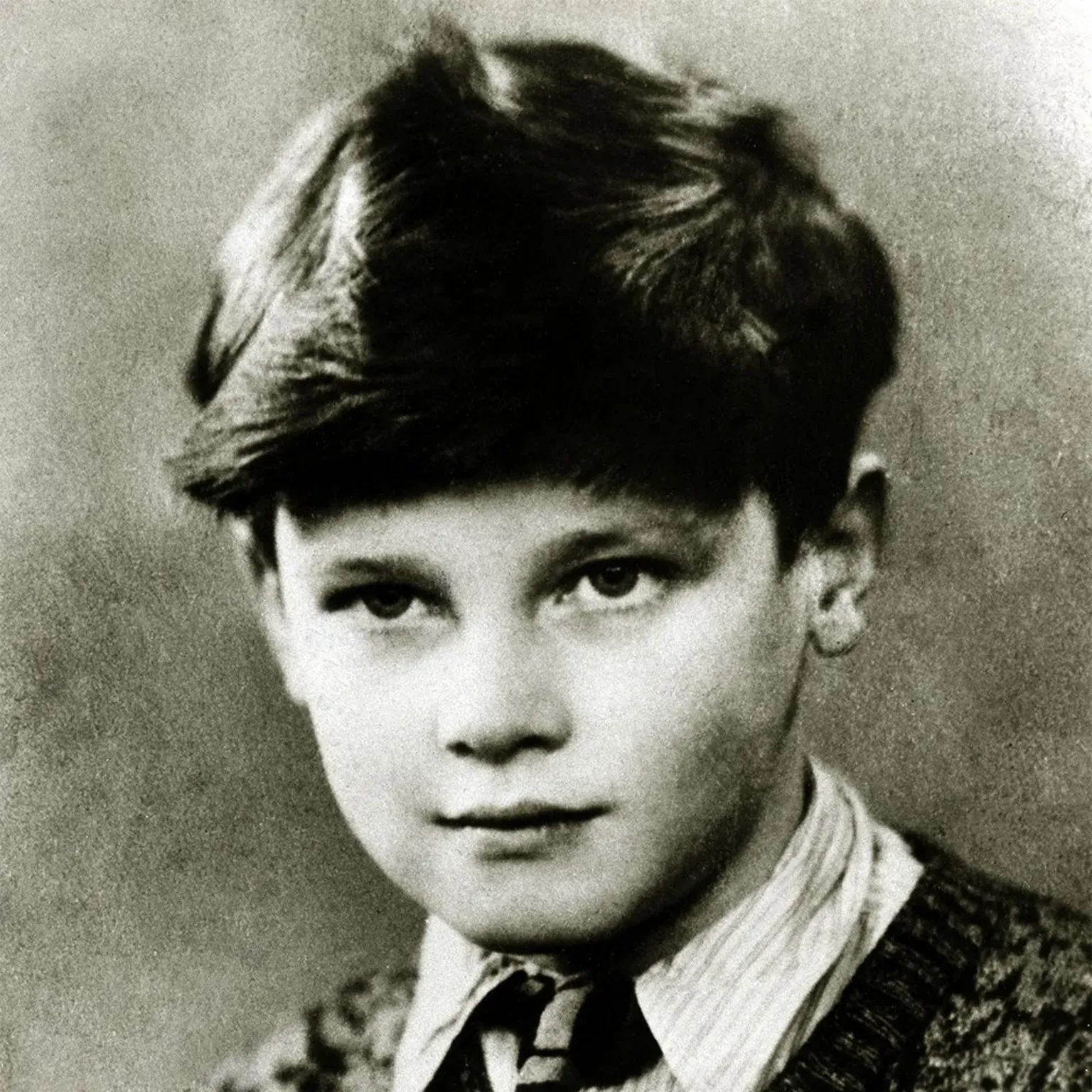A young Sir Roger Moore as a child, wearing a stripy shirt, tie, and knitted jumper.