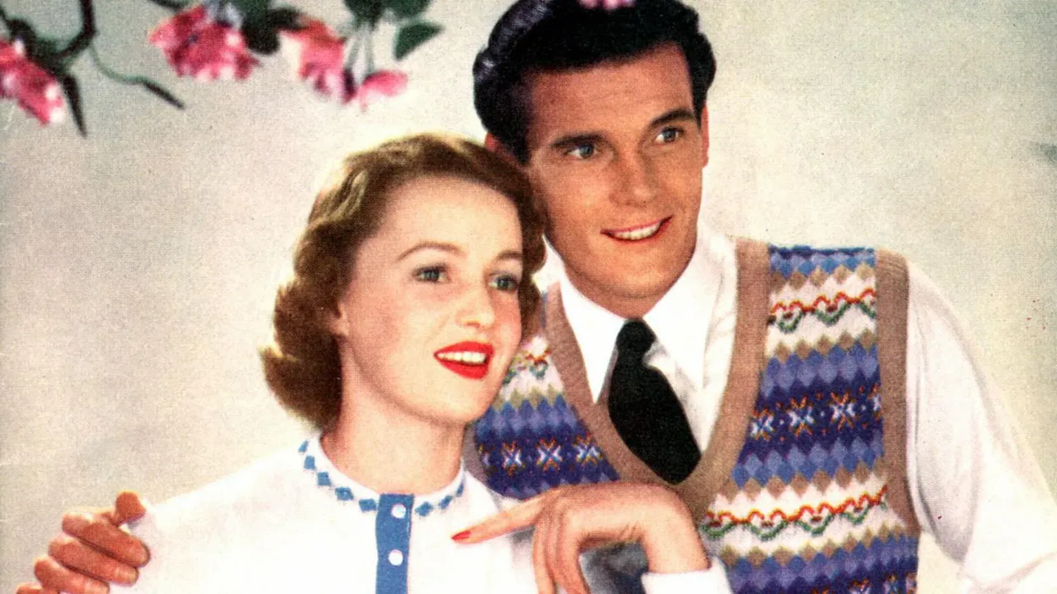 A young Sir Roger Moore modeling a patterned tank top and posing with a woman wearing a white jumper.
