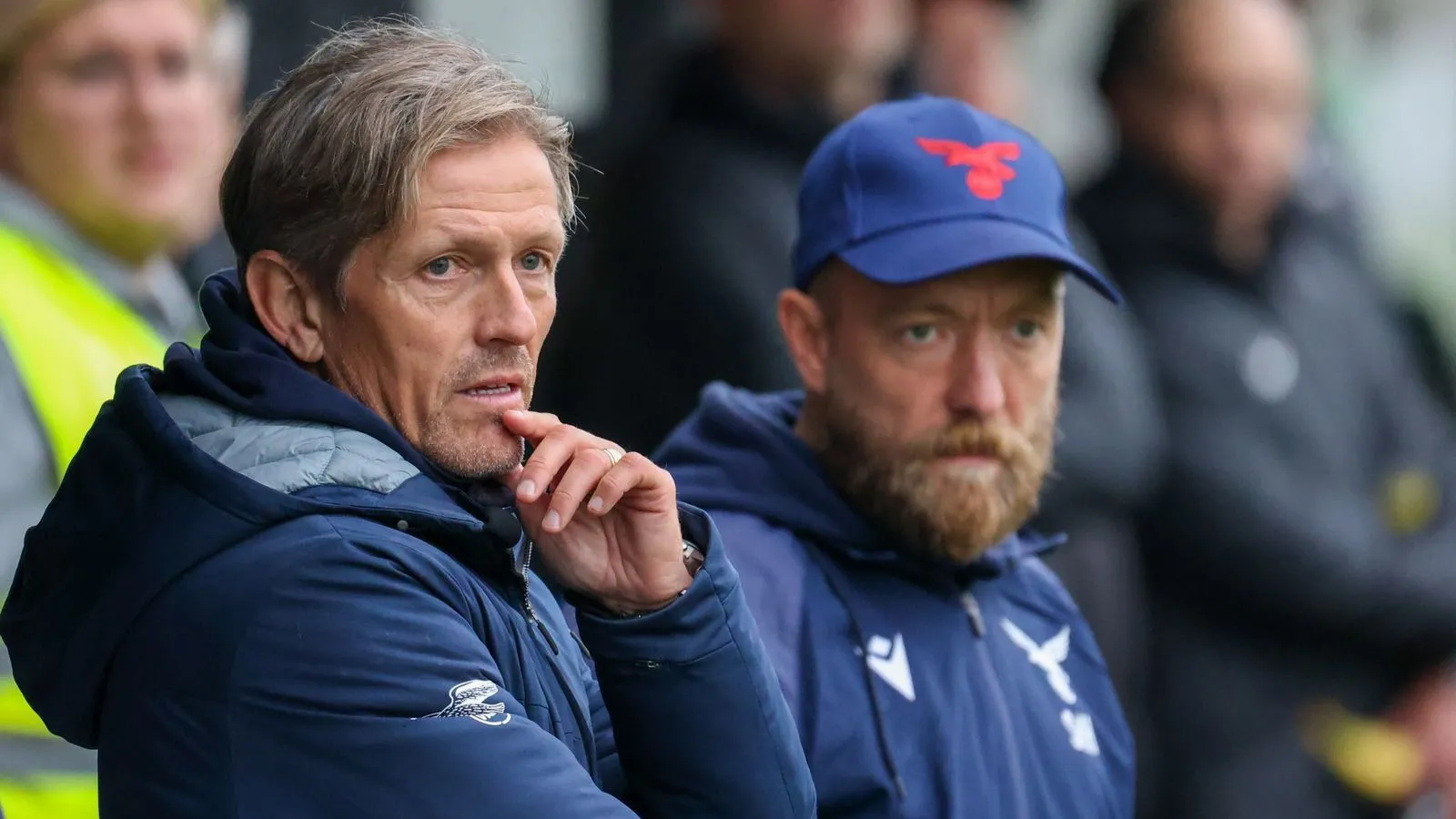Two soccer coaches, one pondering with a finger on his lips, observing the game closely from the sidelines.