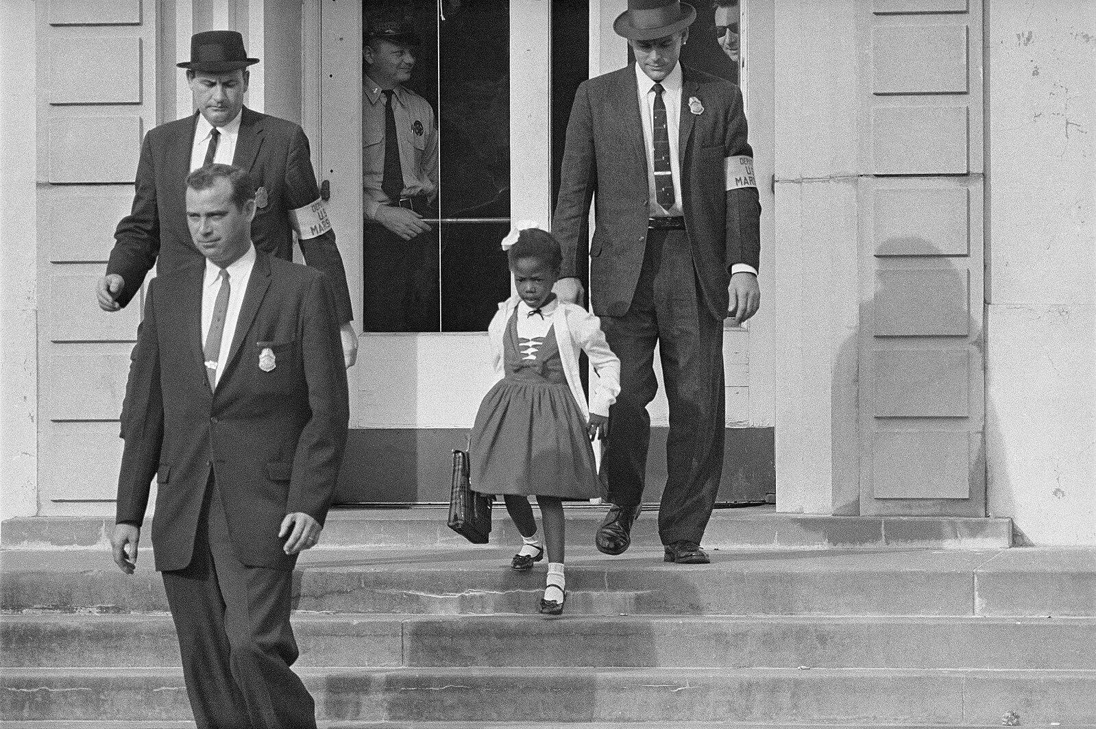 Ruby Bridges, escorted by U.S. marshals to her left and right, walks down the steps in front of a school while carrying a small bag.