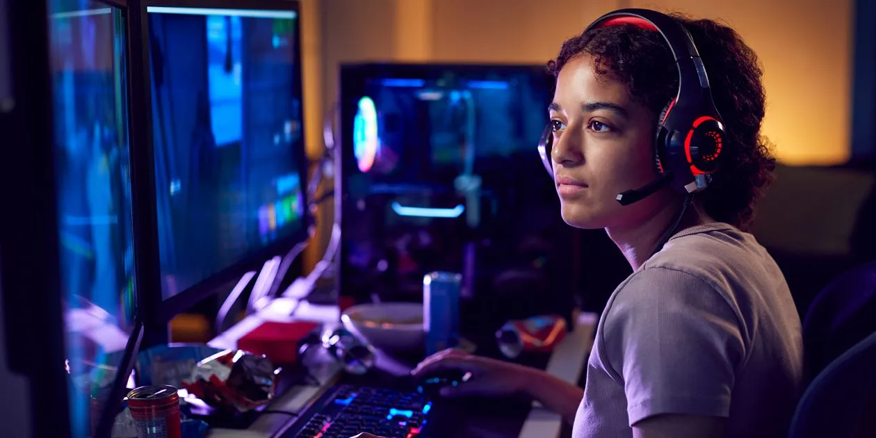 Young woman wearing a headset, gaming using dual computer screens in a dark room