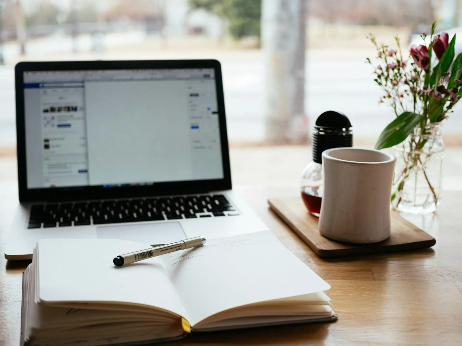 A laptop, notebook with pen, and a coffee mug on a wooden desk, with a blurred outdoor view.
