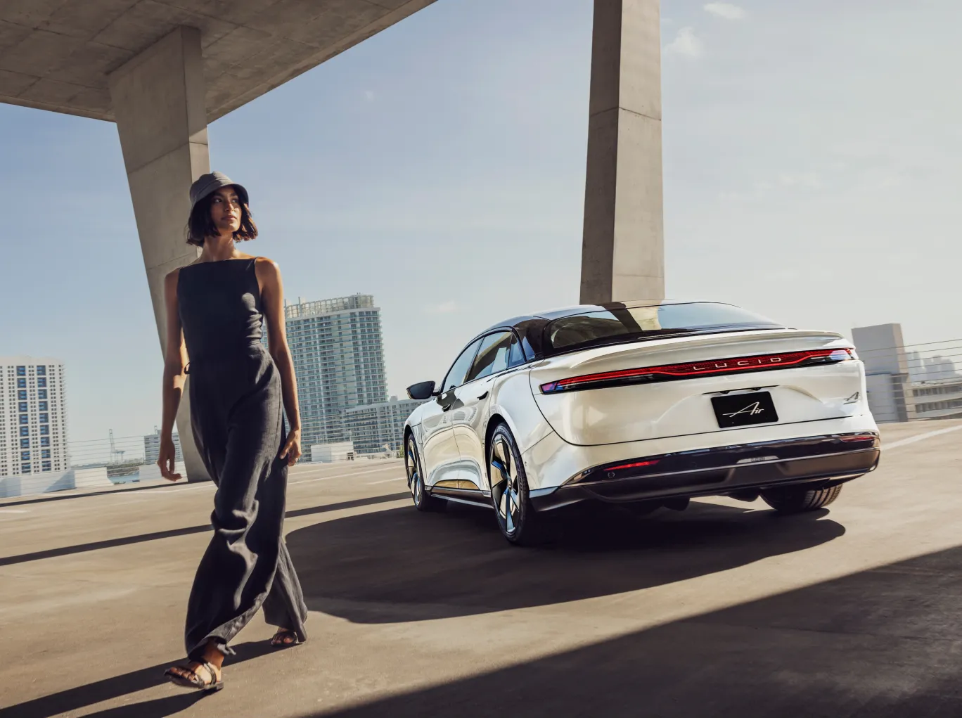 Person in black outfit walking beside a Lucid Air under modern concrete architecture with city skyline.