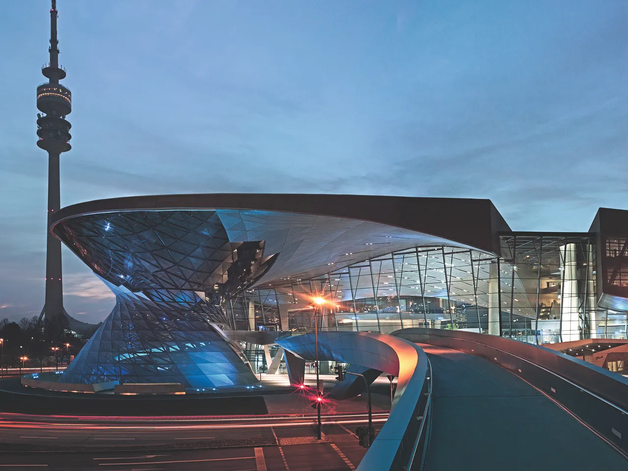 Evening view of BMW Welt with illuminated glass and metal structure, with Munich’s Olympic Tower visible