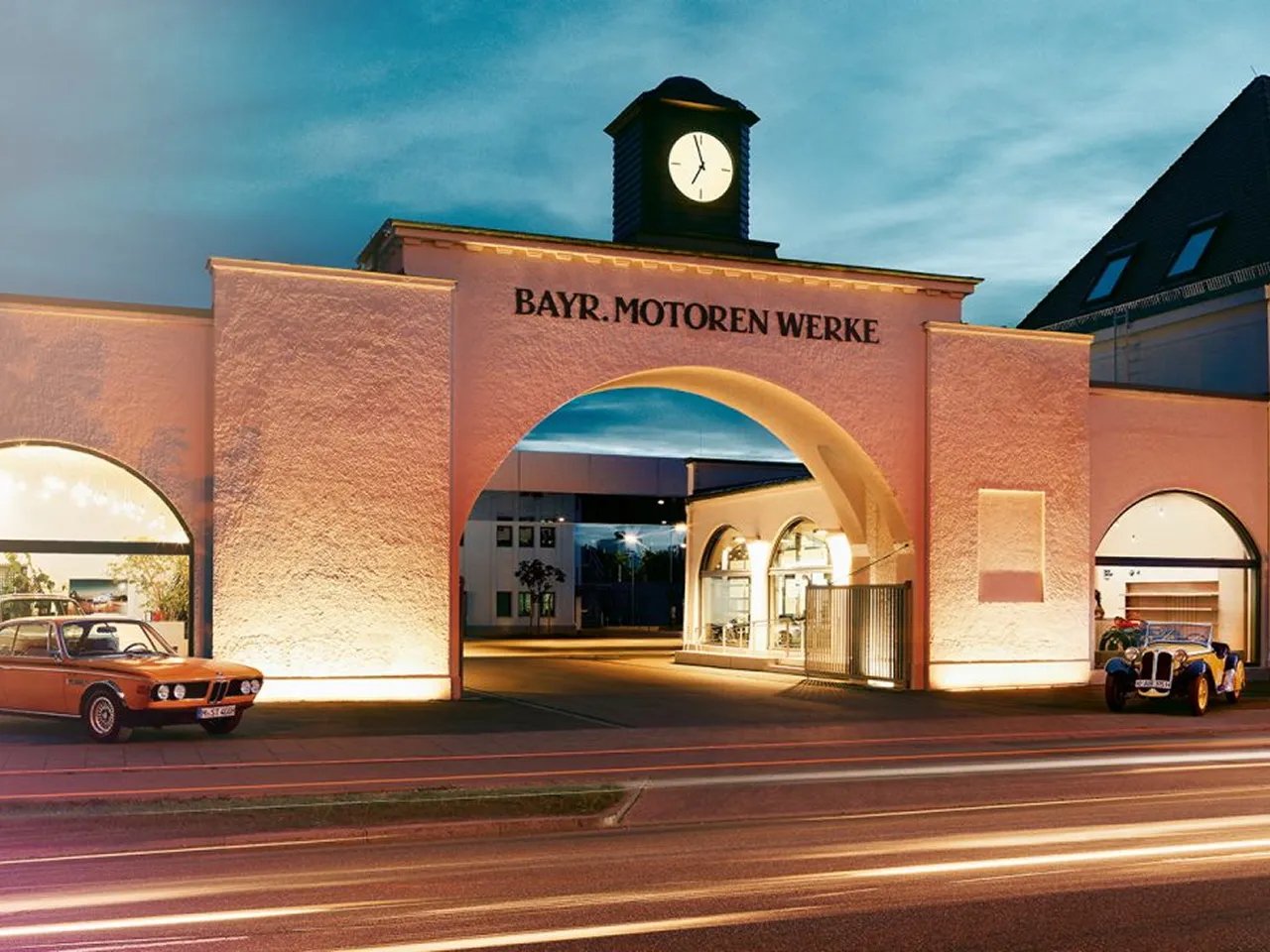 Historic factory gate with clock and the inscription ‘Bayr. Motoren Werke’ at dusk, flanked by two classic BMW vehicles