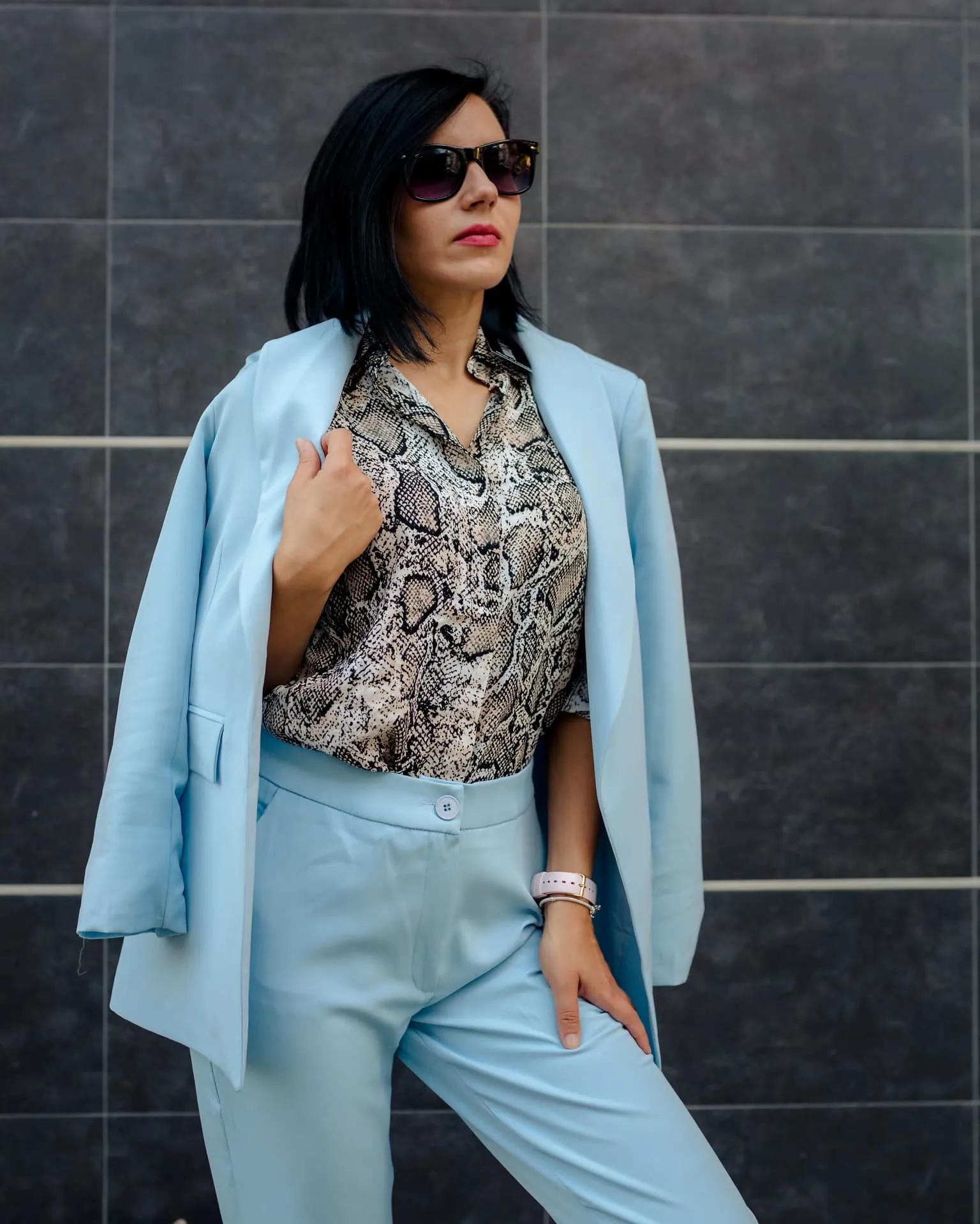 A businesswoman posing in sunglasses, elegant light blue pants, and a tailored coat with a snakeskin-pattern shirt