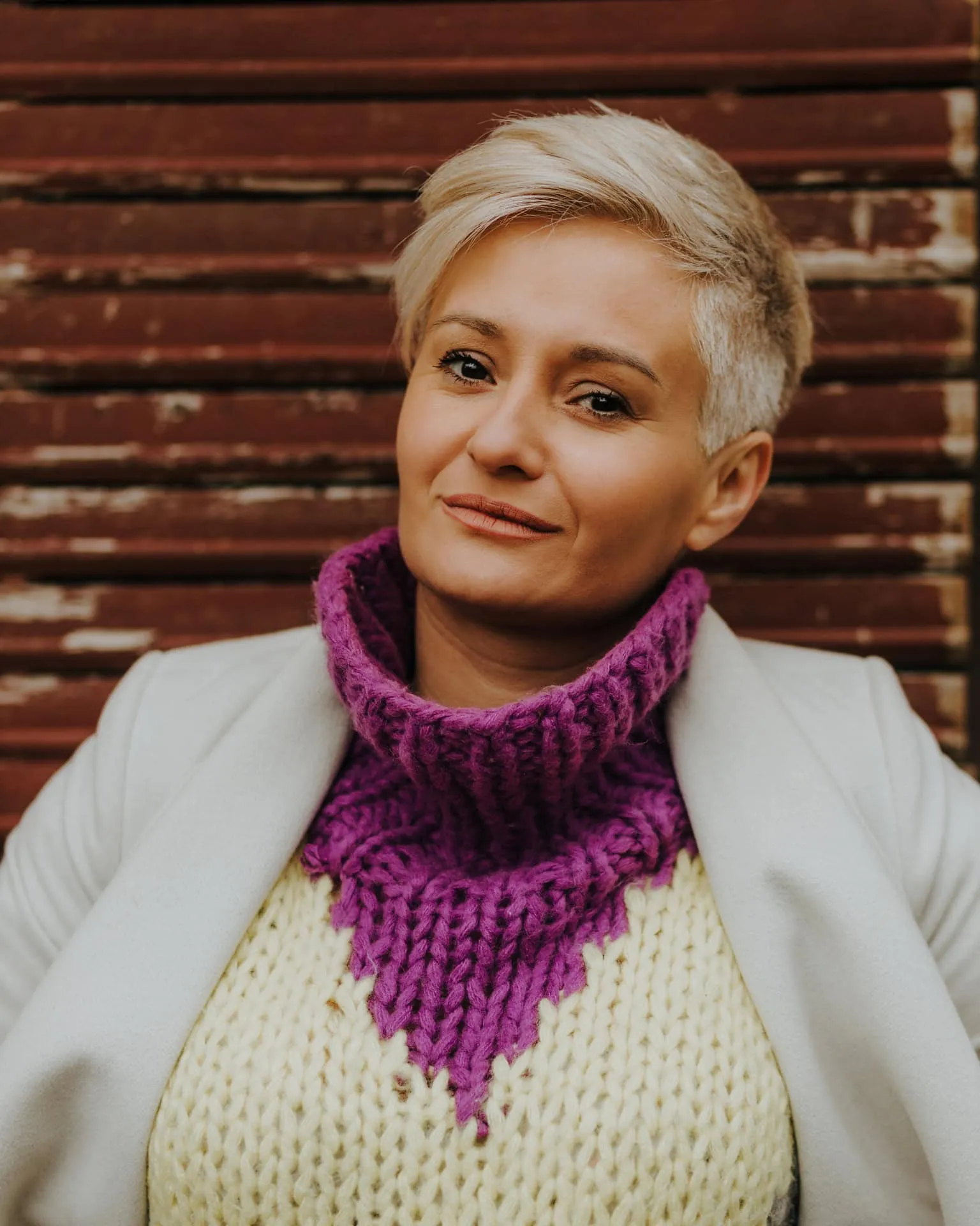 A confidently smiling blonde woman with short hair wearing a handwoven sweater and white jacket, captured in a clean portrait
