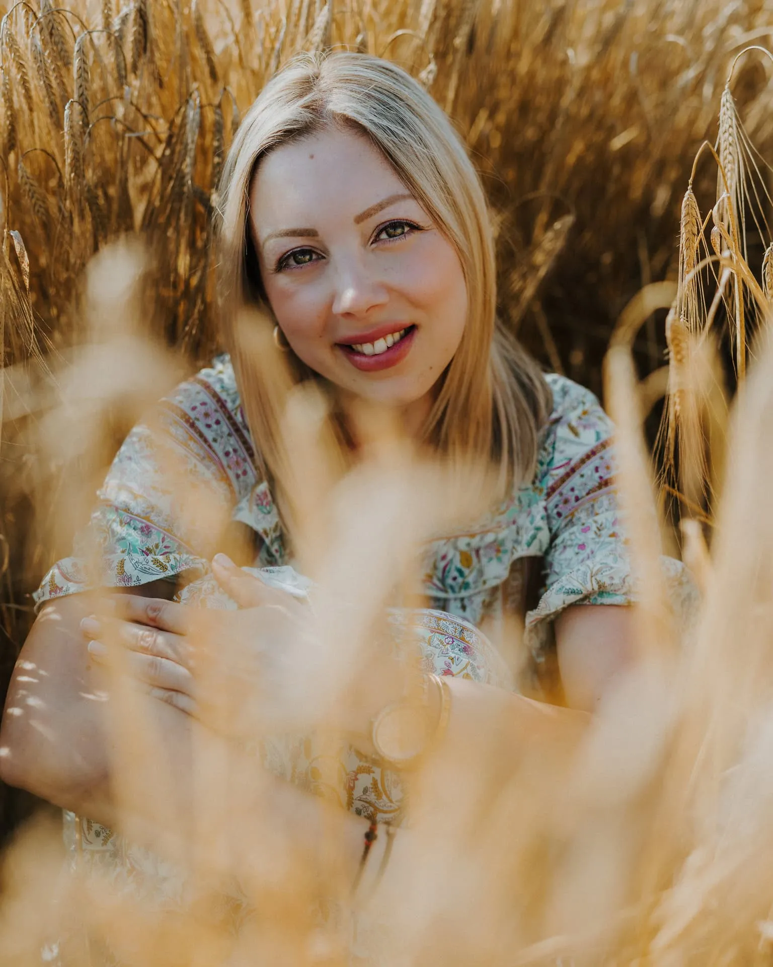 A blonde woman sitting and smiling in a sun-drenched wheat field, surrounded by golden grasses
