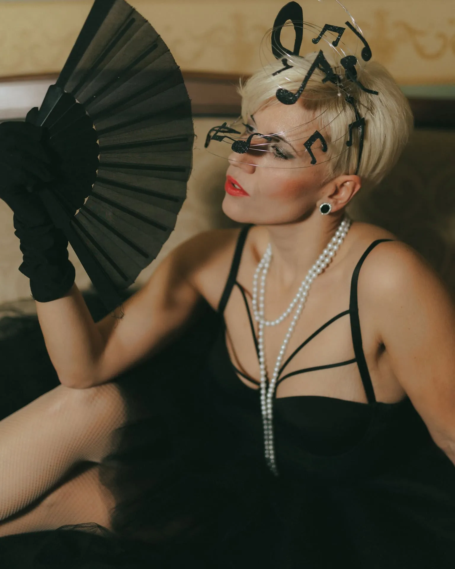 A woman in a vintage-inspired black dress and gloves holding a decorative hand fan, styled in early twentieth-century glamour fashion
