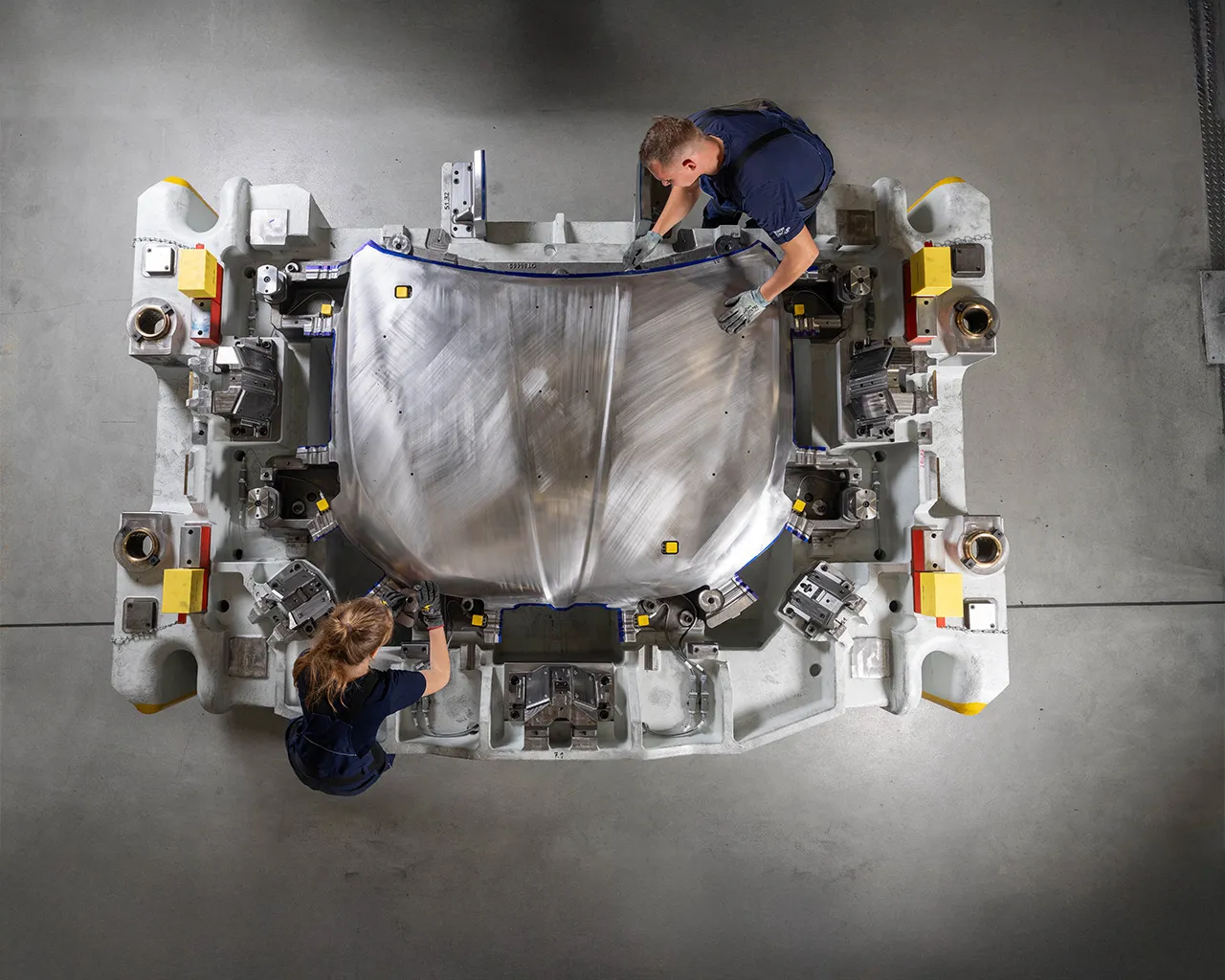Two BMW Group experts using specialised tools on a large metal component in a workshop, viewed from above