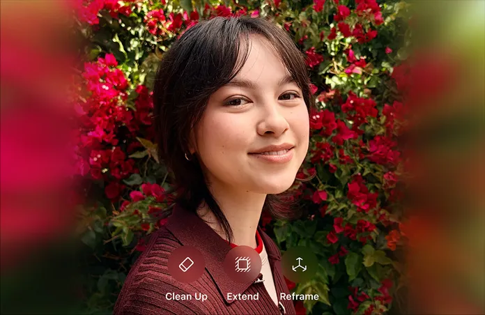 Photo of smiling person with brown hair, wearing a brown sweater, surrounded by red flowers, AI photo editing demo with Clean Up, Extend, Reframe options.
