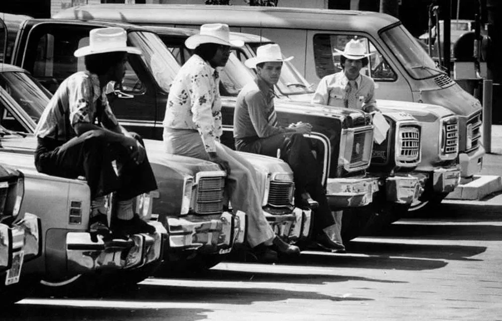Group of car salesmen sitting around vehicles at a dealership lot in late 1970s California