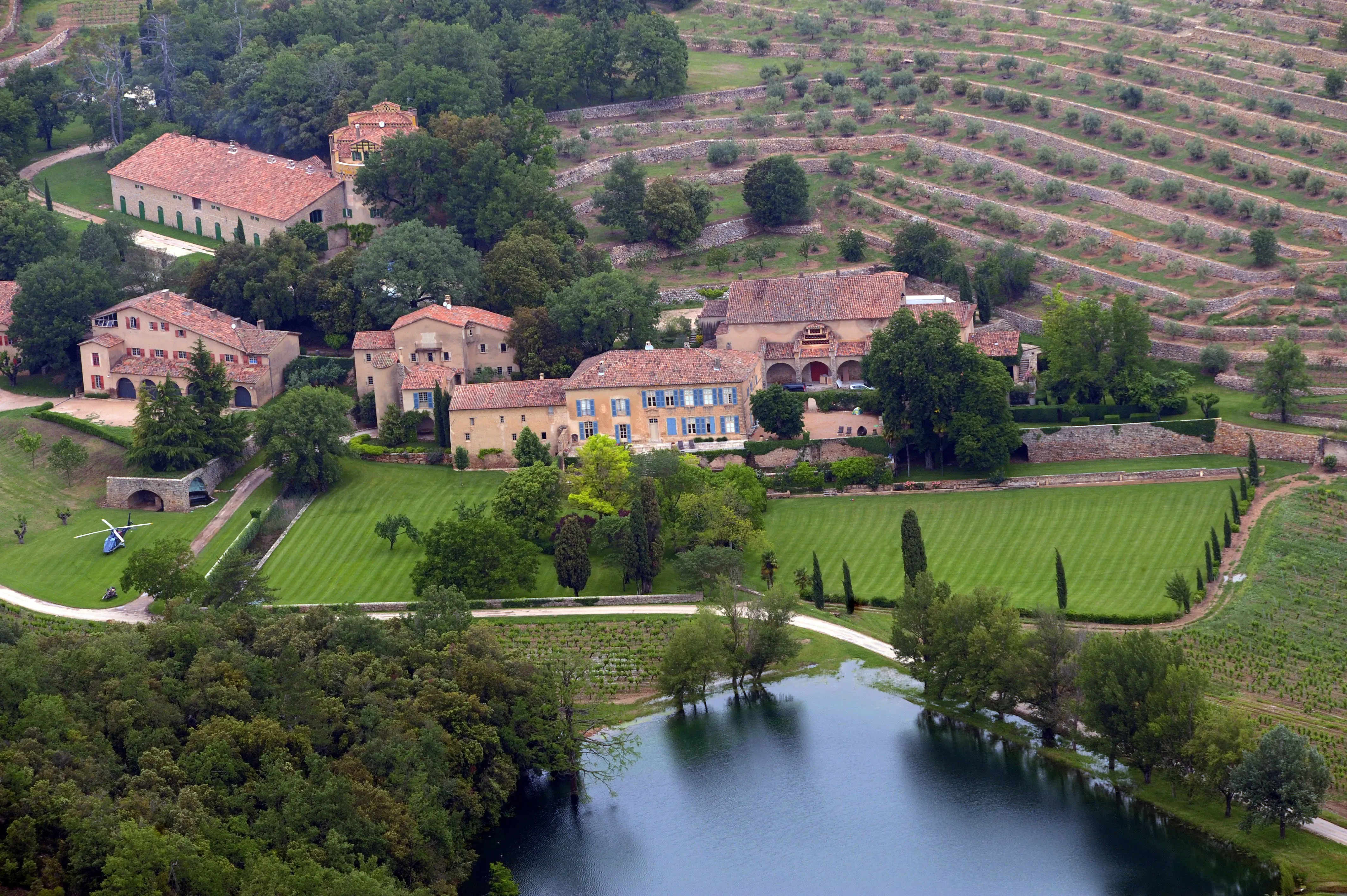 Aerial view of Chateau Miraval, a large estate with several buildings and surrounding vineyards, with a lake in the foreground.
