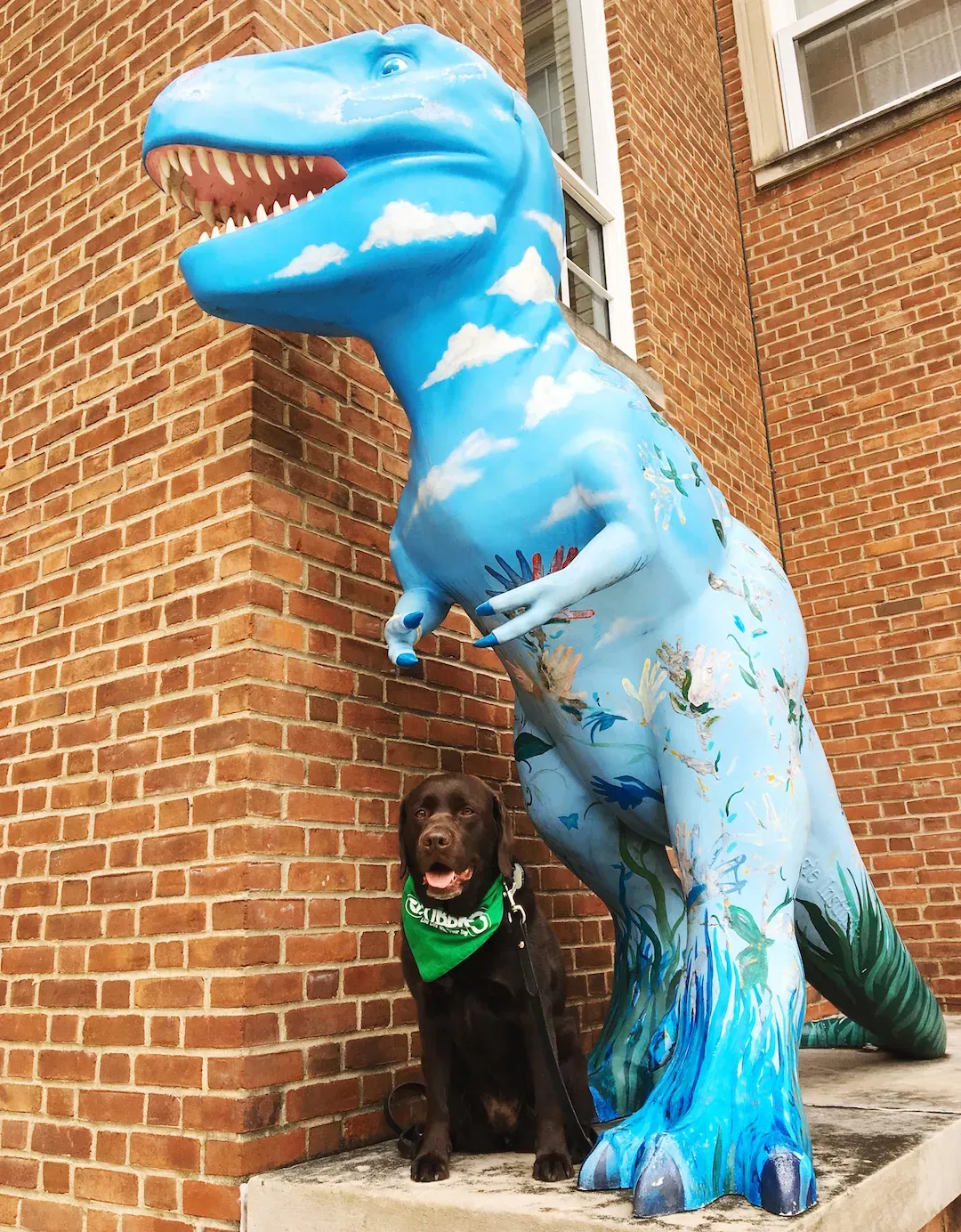 Black lab mix wearing a branded bandana sitting next to a dinosaur statue